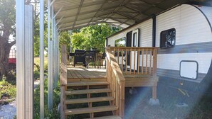 A covered outdoor deck with a wooden staircase leading to a covered area, a barbecue grill, and a dining table with chairs.