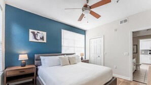 A bedroom with a bed, nightstands, a ceiling fan, a window with blinds, and a door leading to a bathroom. - Landing | Modern 1BD, Gym, Pool (Orlando)