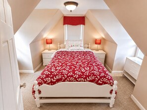 A bedroom with a sloped ceiling, a bed with a red leaf-patterned bedspread, two bedside tables with lamps, and a window with blinds.