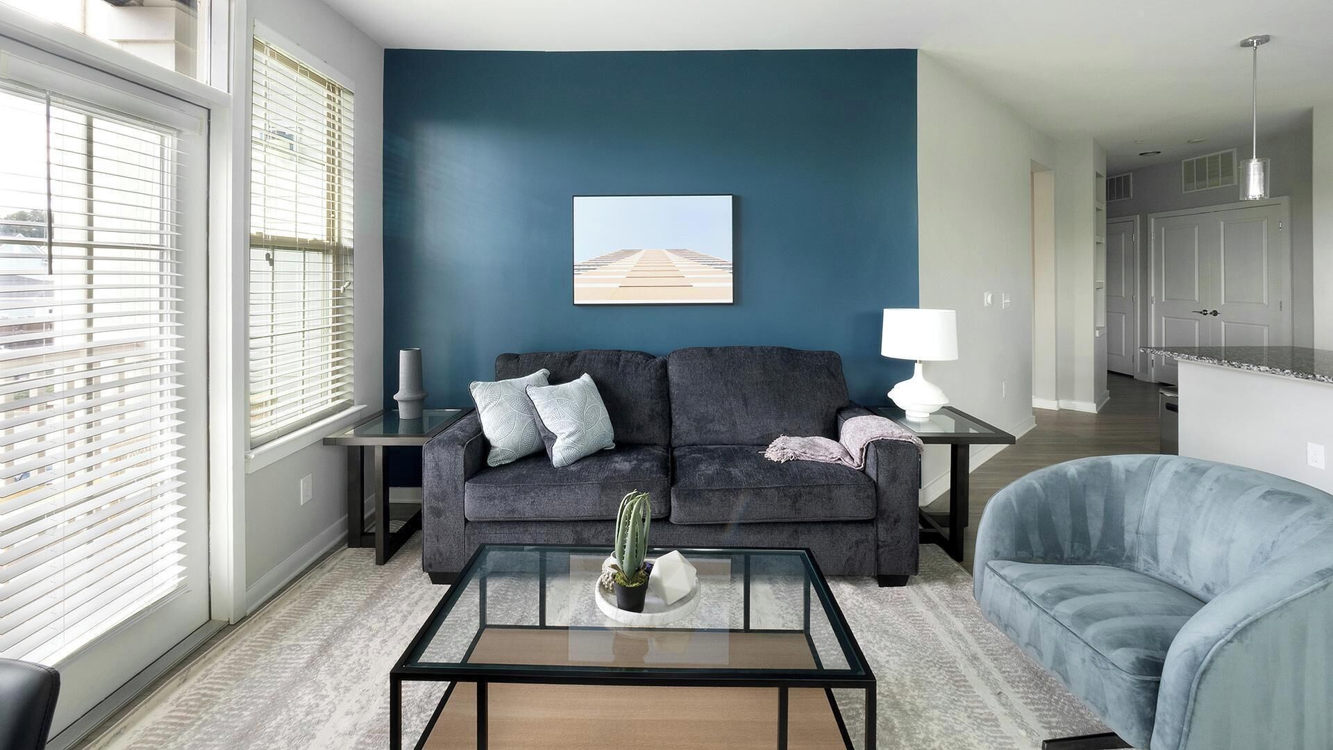 Una sala moderna con una pared azul oscuro, un sofá gris, un sillón azul, una mesa de centro de vidrio y vistas de la ciudad a través de amplios ventanales.