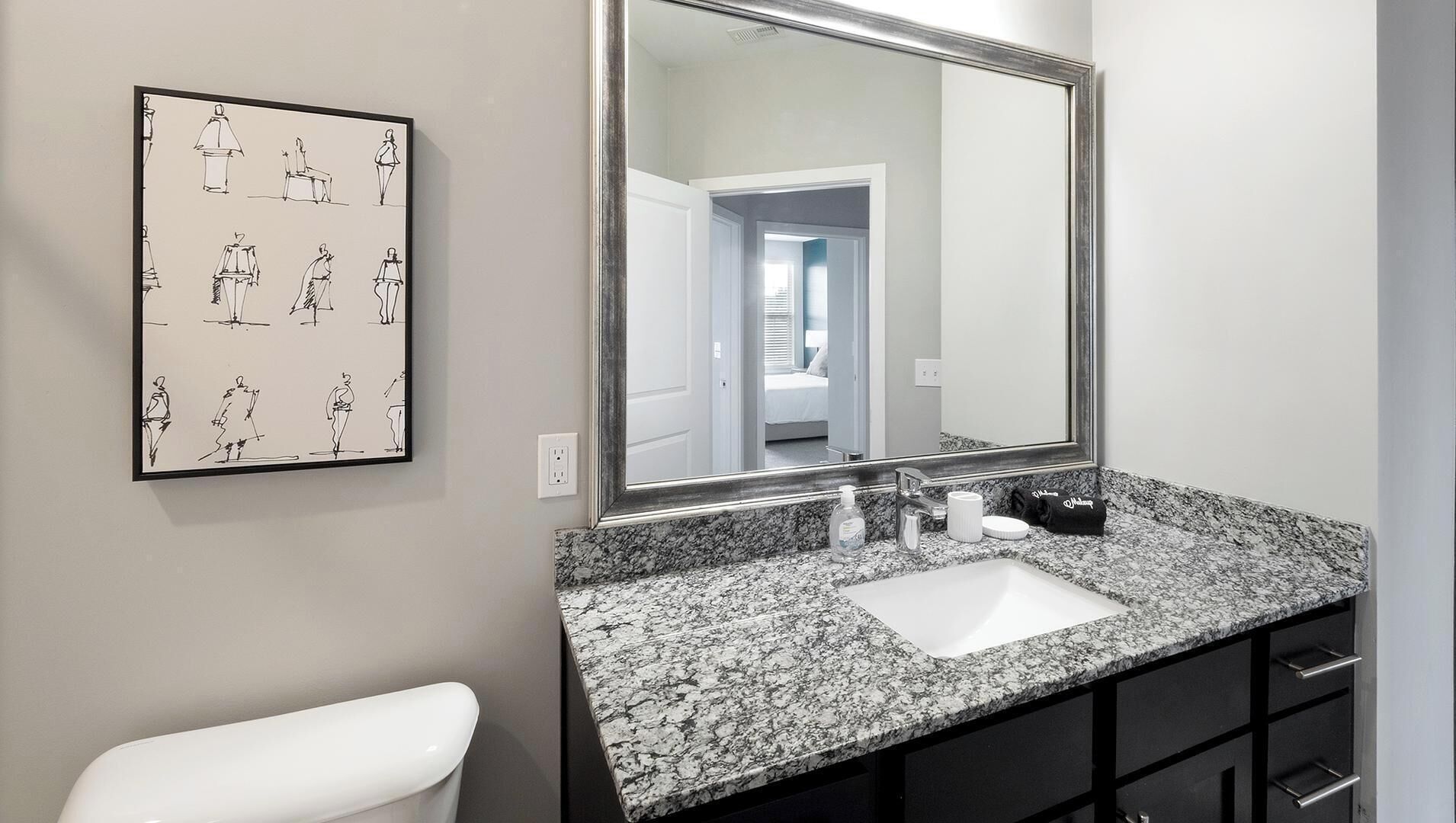 A bathroom with a granite countertop, a white sink, a large mirror, and a wall-mounted artwork depicting various figures.