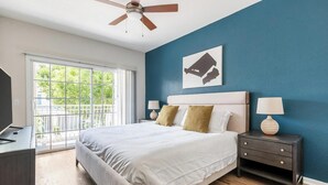A bedroom with a bed, nightstands, a ceiling fan, a window with a view, and a wall with a painting. - Landing | Stylish 2BD, Gym, Pool (Orlando)