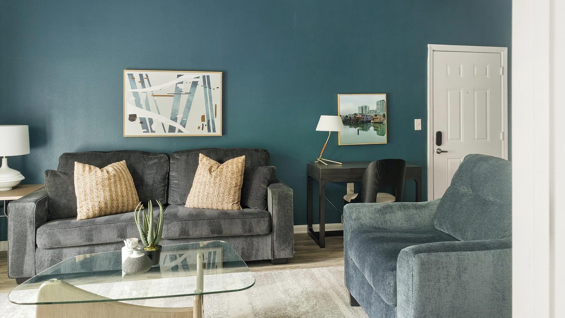 Una sala de estar con un sofá gris, un sillón azul y una mesa de centro de vidrio.