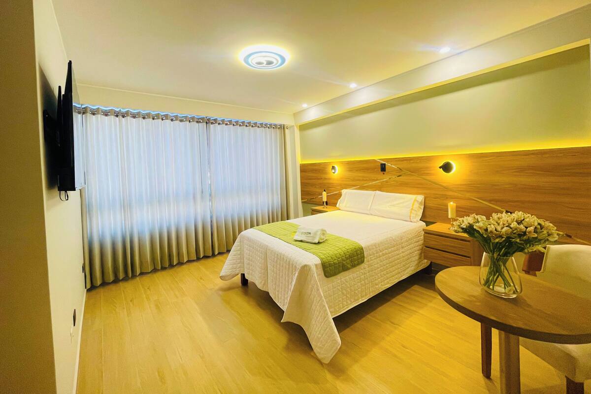 A hotel room with a bed, bedside table, lamp, and a small table with a vase of flowers.