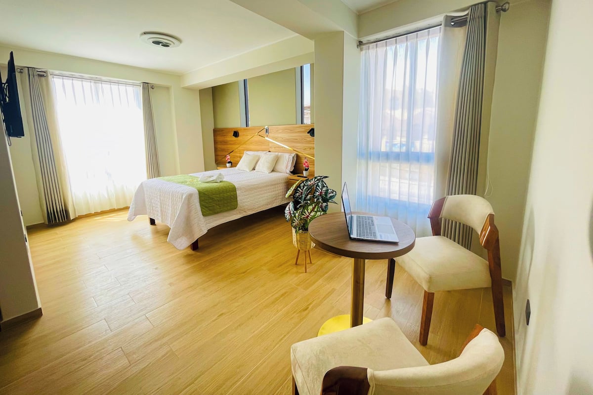 A hotel room with a bed, a desk with a laptop, a chair, and a potted plant.