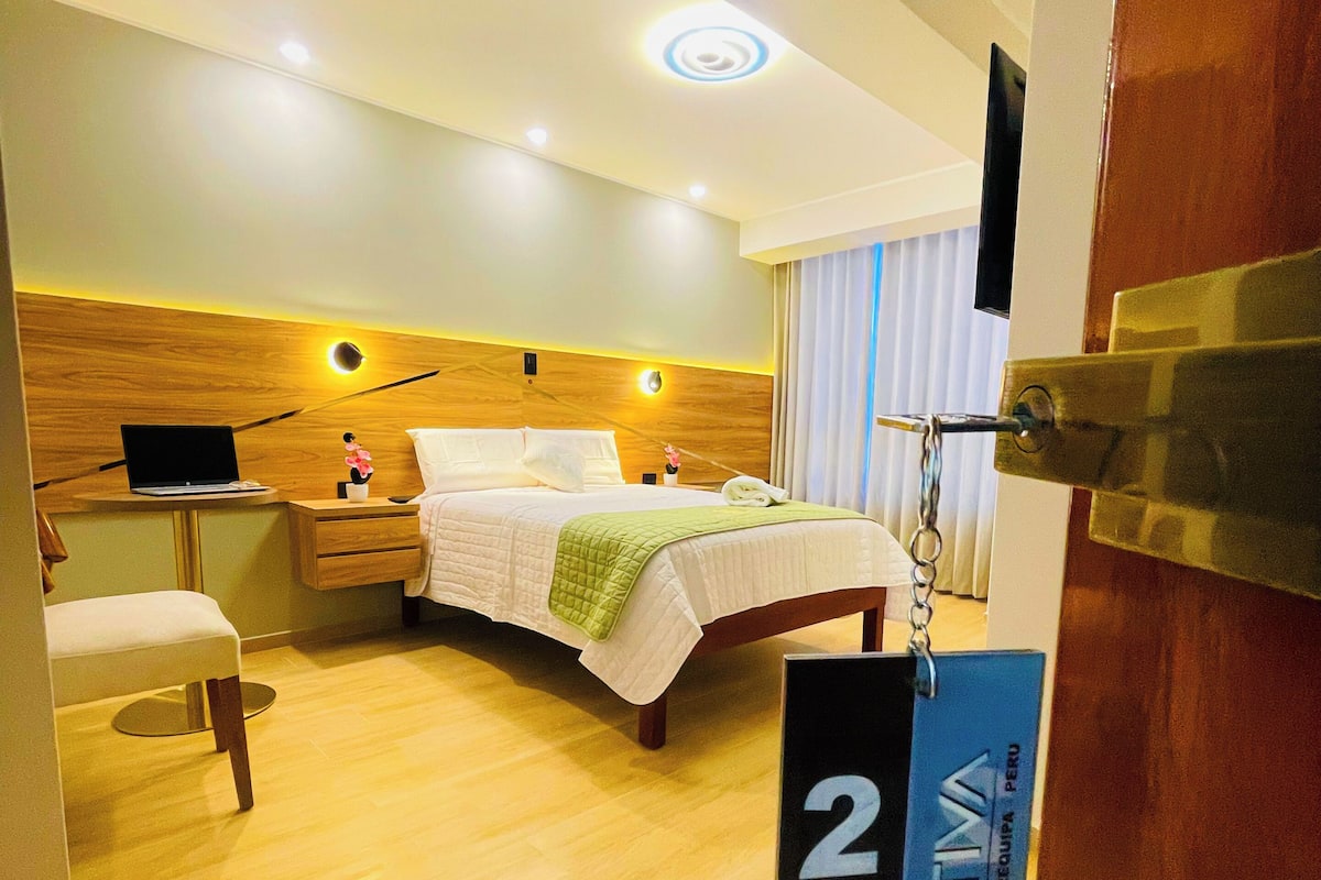 A hotel room with a bed, bedside table, lamp, and a TV mounted on the wall.