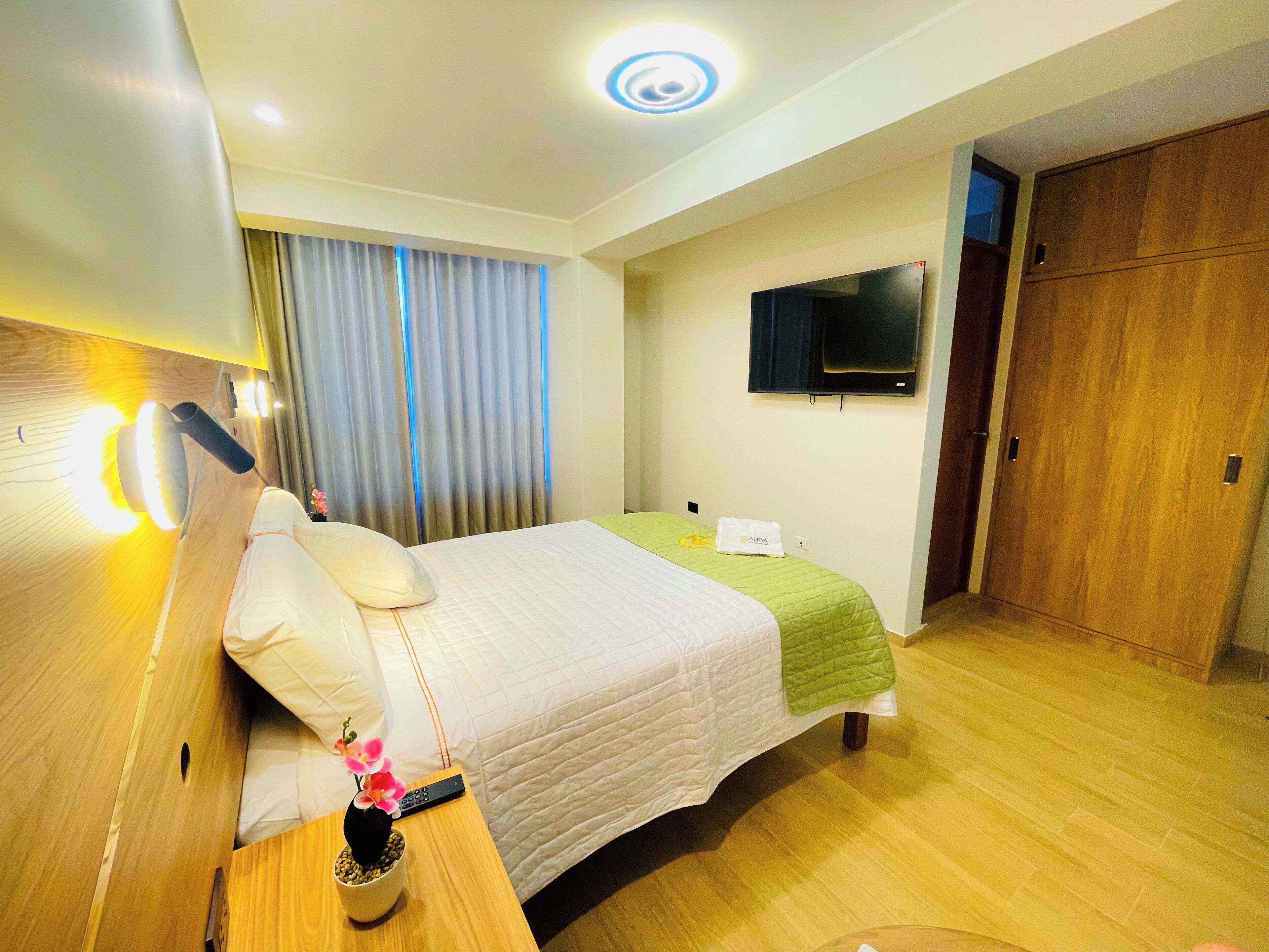 A hotel room with a bed, a TV mounted on the wall, a wooden wardrobe, and a nightstand with a remote control and a vase with flowers.