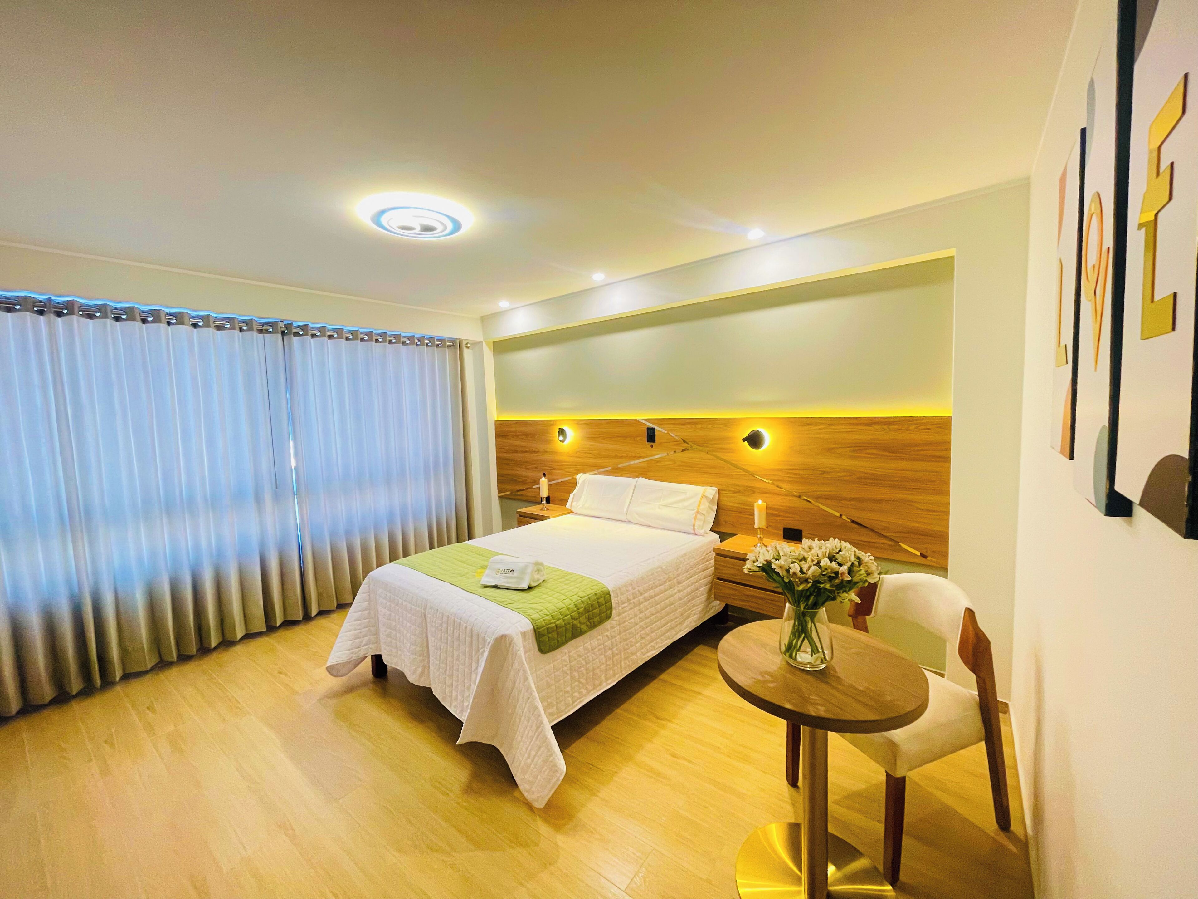 Habitación de hotel con cama, mesita de noche con un florero, una silla y una pequeña mesa.
