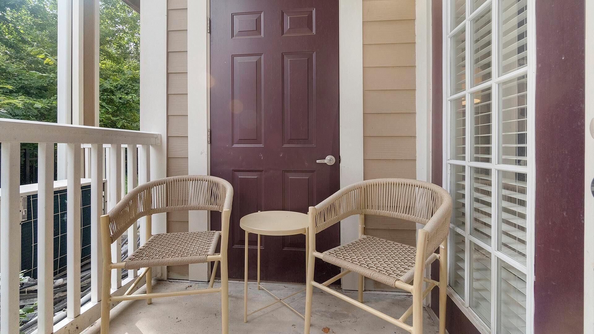 Une véranda avec deux fauteuils en osier et une petite table ronde, une porte marron foncé et une fenêtre avec des stores.