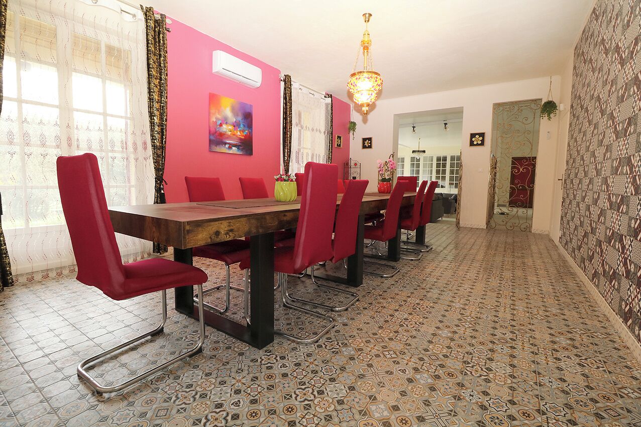 Un espace repas avec une longue table en bois et des chaises rouges, un lustre, des carreaux de sol à motifs et un mur rose orné d’un tableau.