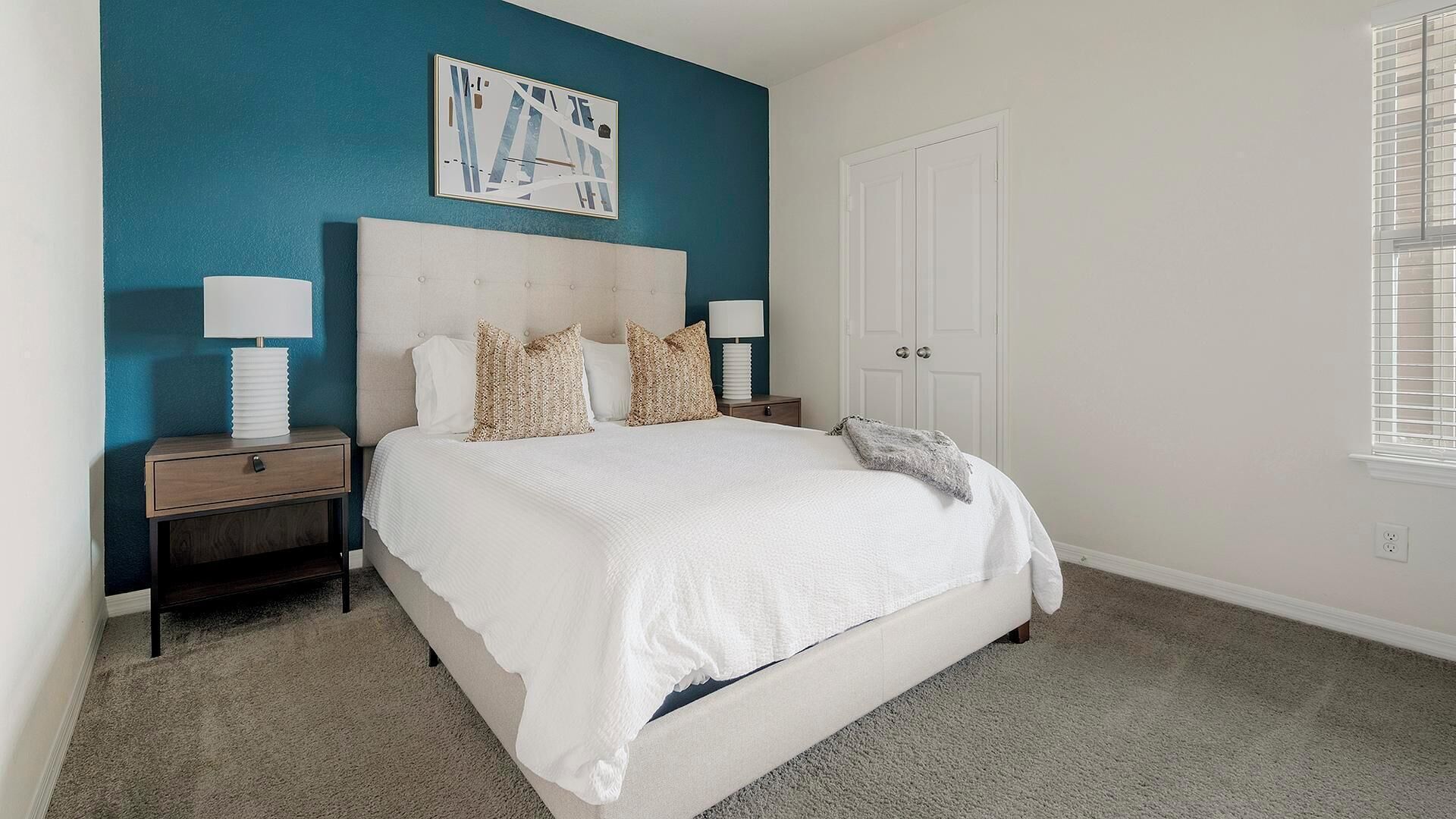 A bedroom with a large bed, two bedside tables, a nightstand, a door, and a window with blinds.