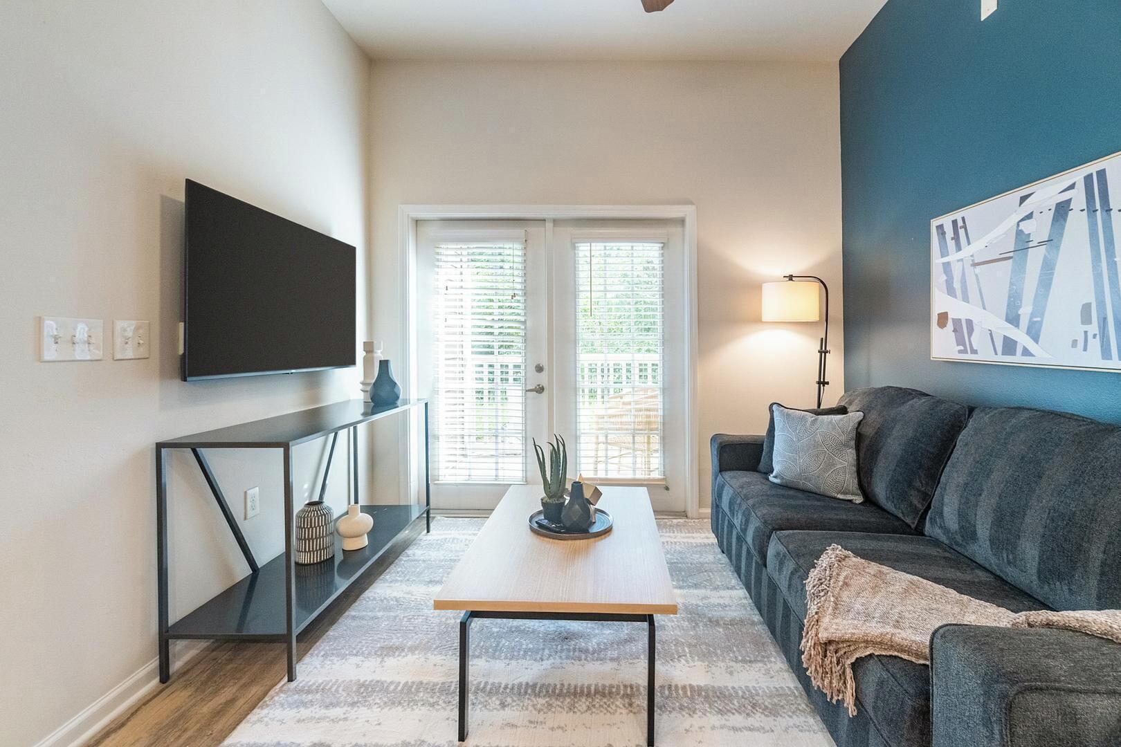 A living room with a sofa, a coffee table, a TV mounted on the wall, and a door with a view.