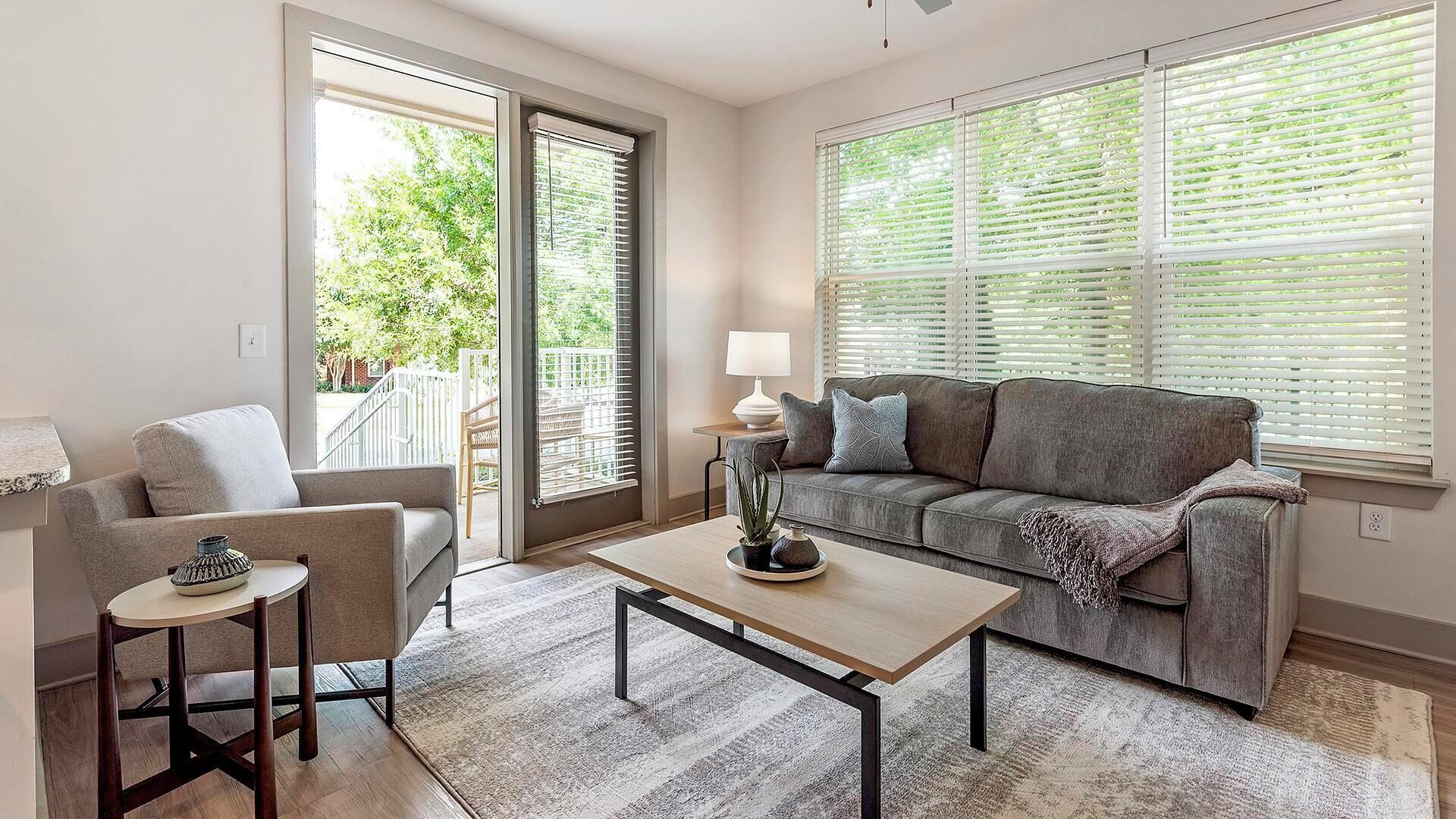 A living room with a sofa, a coffee table, and a chair. There is a window with blinds and a view of greenery outside.