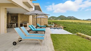 Un área de descanso exterior moderna con sillones azules y sombrillas a rayas, piscina y vistas a colinas verdes.
