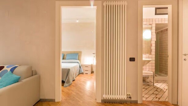 A modern hotel room with a wooden floor, a bed with blue bedding, a white radiator, and a bathroom visible through an open door. - Charm and comfort at a stone's throw from the Arena (Verona)