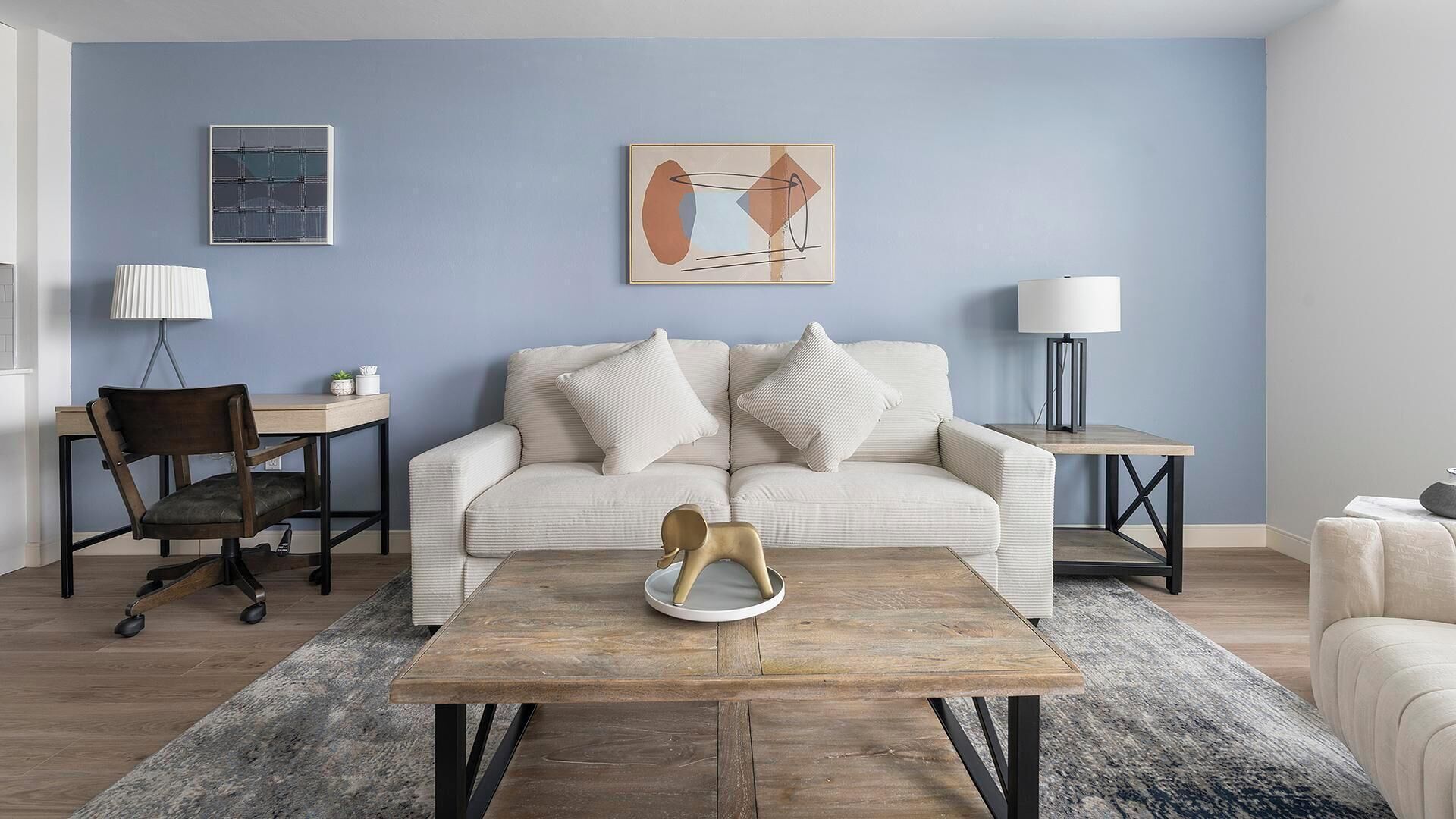 A modern living room with a light blue wall, a beige sofa with two pillows, a wooden coffee table, a desk with a chair, and two floor lamps.