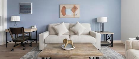 A modern living room with a light blue wall, a beige sofa with two pillows, a wooden coffee table, a desk with a chair, and two floor lamps.