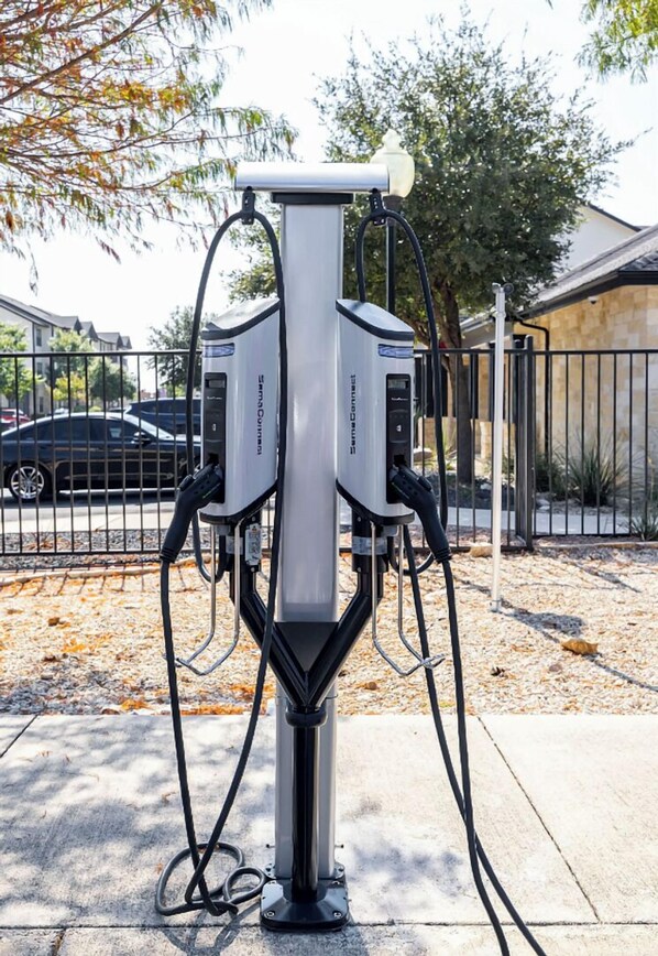Une borne de recharge dotée de deux unités, reliées par des câbles, installée à l’extérieur, avec une clôture et des voitures garées en arrière-plan.