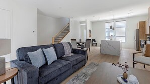 Ett modernt vardagsrum med en grå soffa, en matplats och ett kök med träskåp.