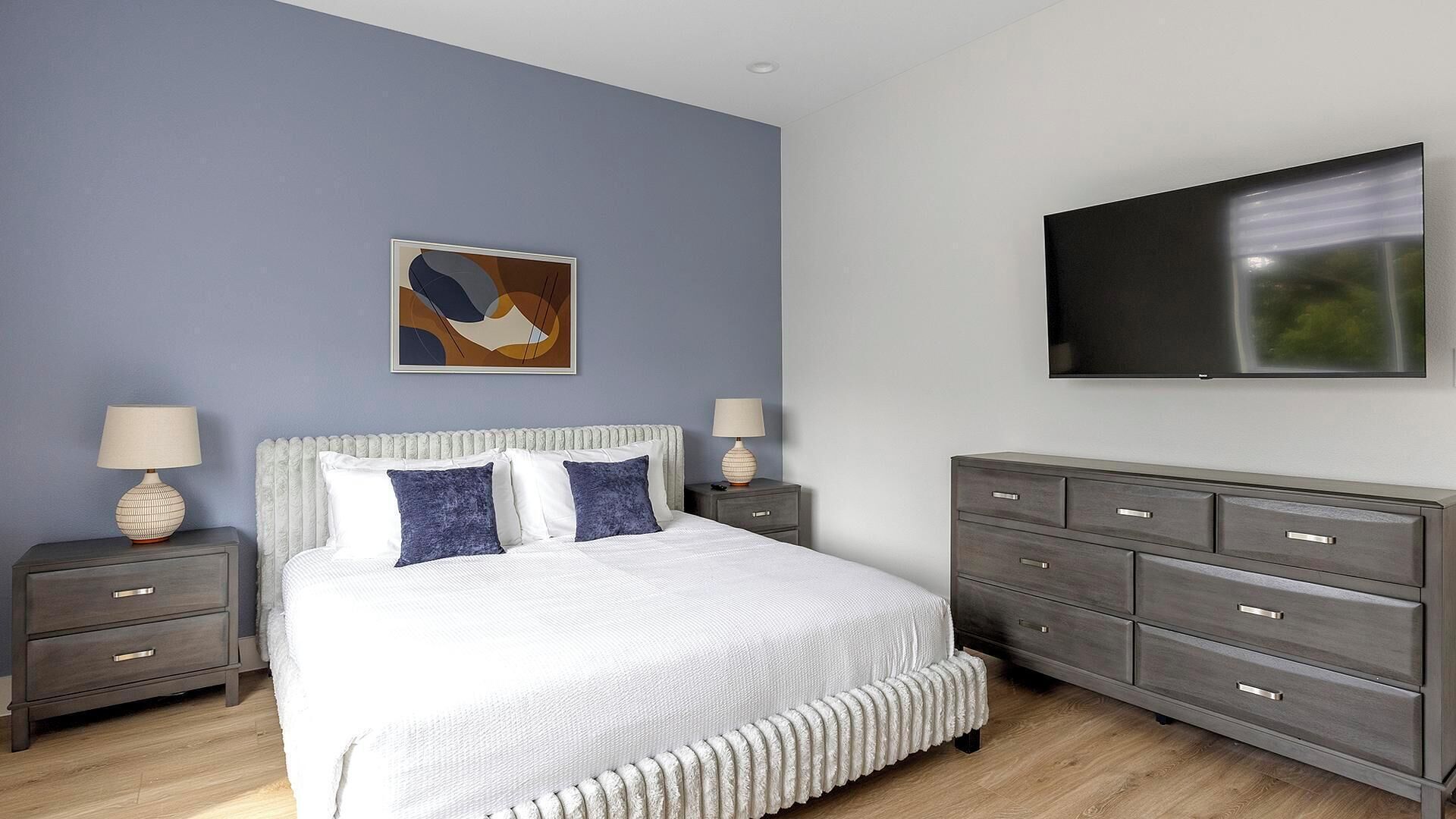 A bedroom with a bed, two bedside tables, a dresser, a television, and a wall-mounted artwork.