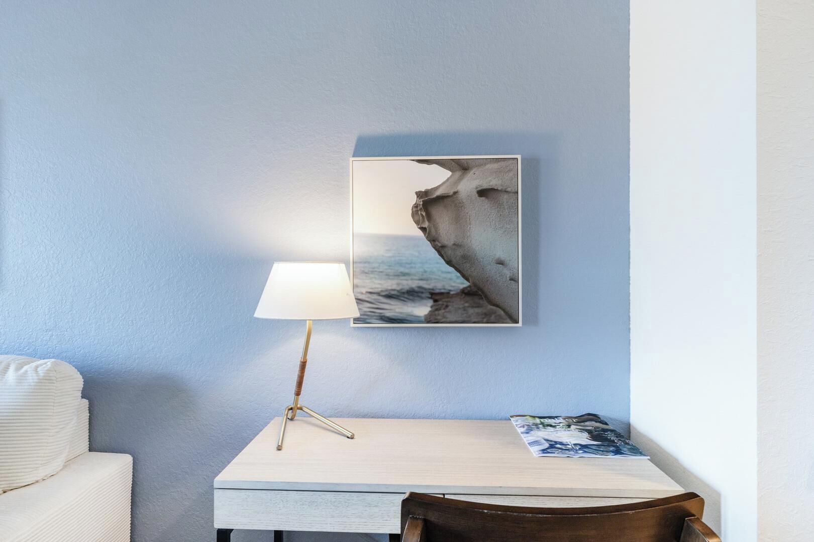 A room with a white table, a lamp, a framed abstract painting, and a chair.
