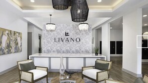 Un área de recepción con un mostrador blanco, dos sillas de mimbre, una lámpara de araña moderna y una pared con el letrero 'LIVANO'.