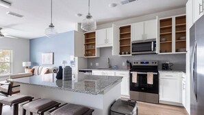Una cocina moderna con gabinetes blancos, una isla central con taburetes de bar, electrodomésticos de acero inoxidable e iluminación colgante.