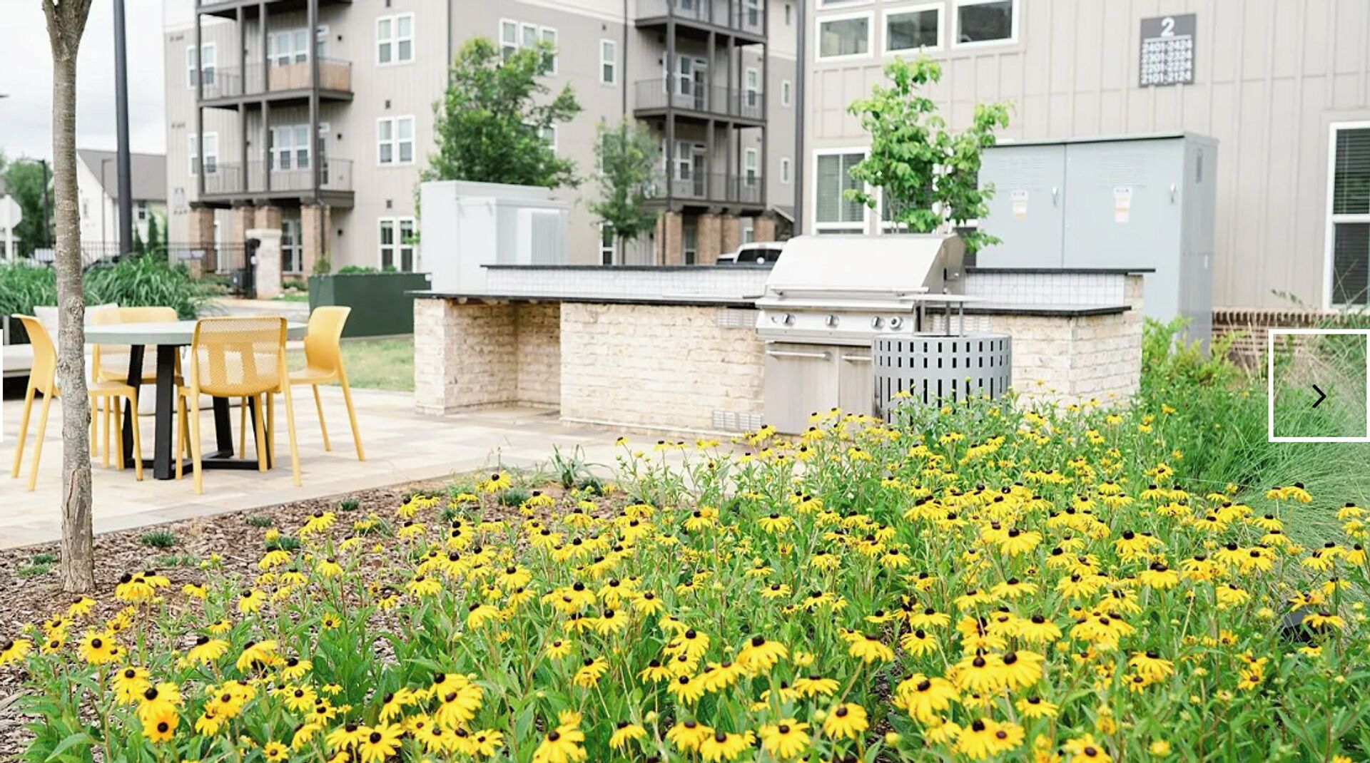 An outdoor patio with a grill, yellow chairs, and a flower bed with yellow flowers.
