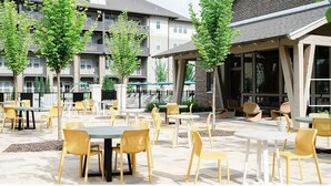 An outdoor patio area with multiple tables and chairs, surrounded by trees and a building with balconies.