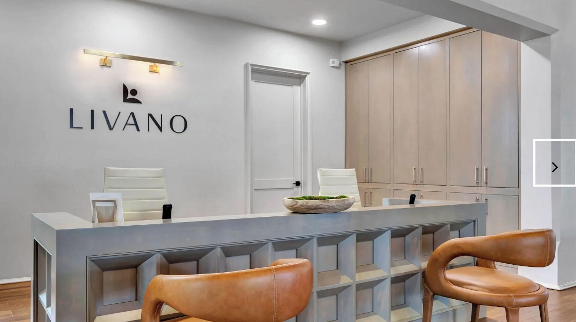 Un comptoir d’accueil moderne au design minimaliste, un grand miroir et une enseigne indiquant « LIVANO ». Deux fauteuils en cuir sont placés devant le comptoir.