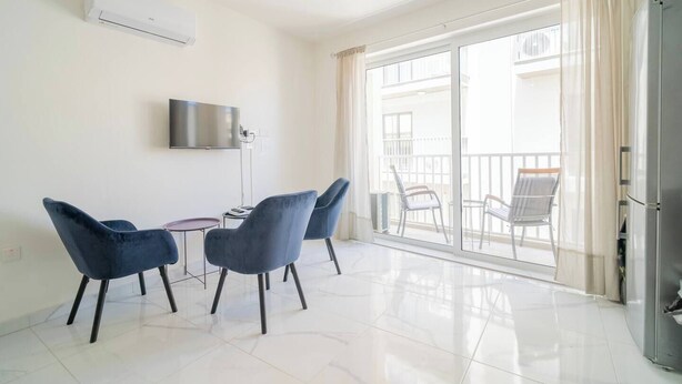 A modern living room with a flat-screen TV, two blue chairs, a small table, and a sliding glass door leading to an outdoor balcony with chairs. - Cozy Apartment with Balcony in Xlendi Bay, Gozo (Munxar)