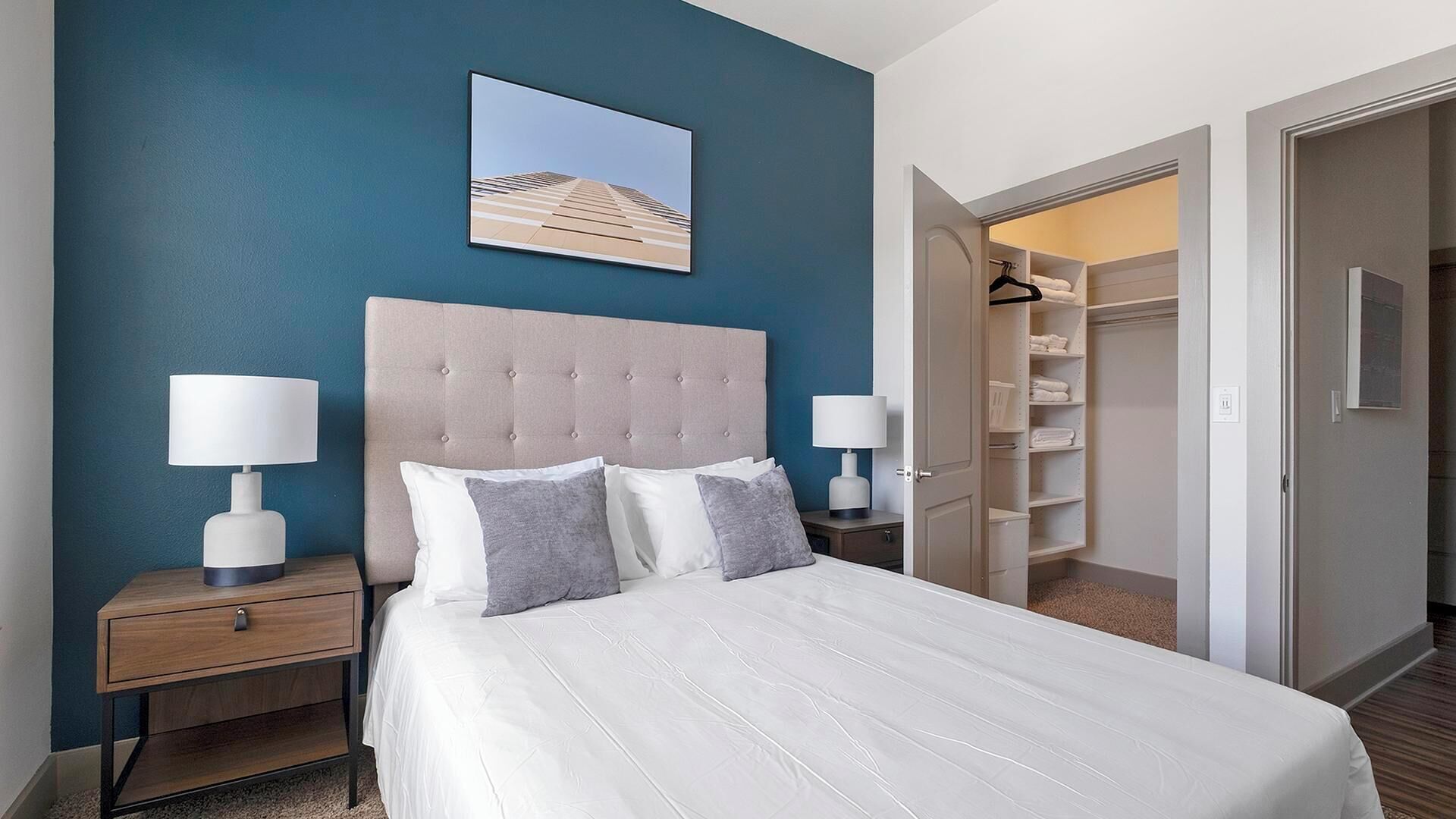 A hotel room with a large bed, two bedside tables with lamps, a headboard, a wardrobe, and a framed picture on the wall.
