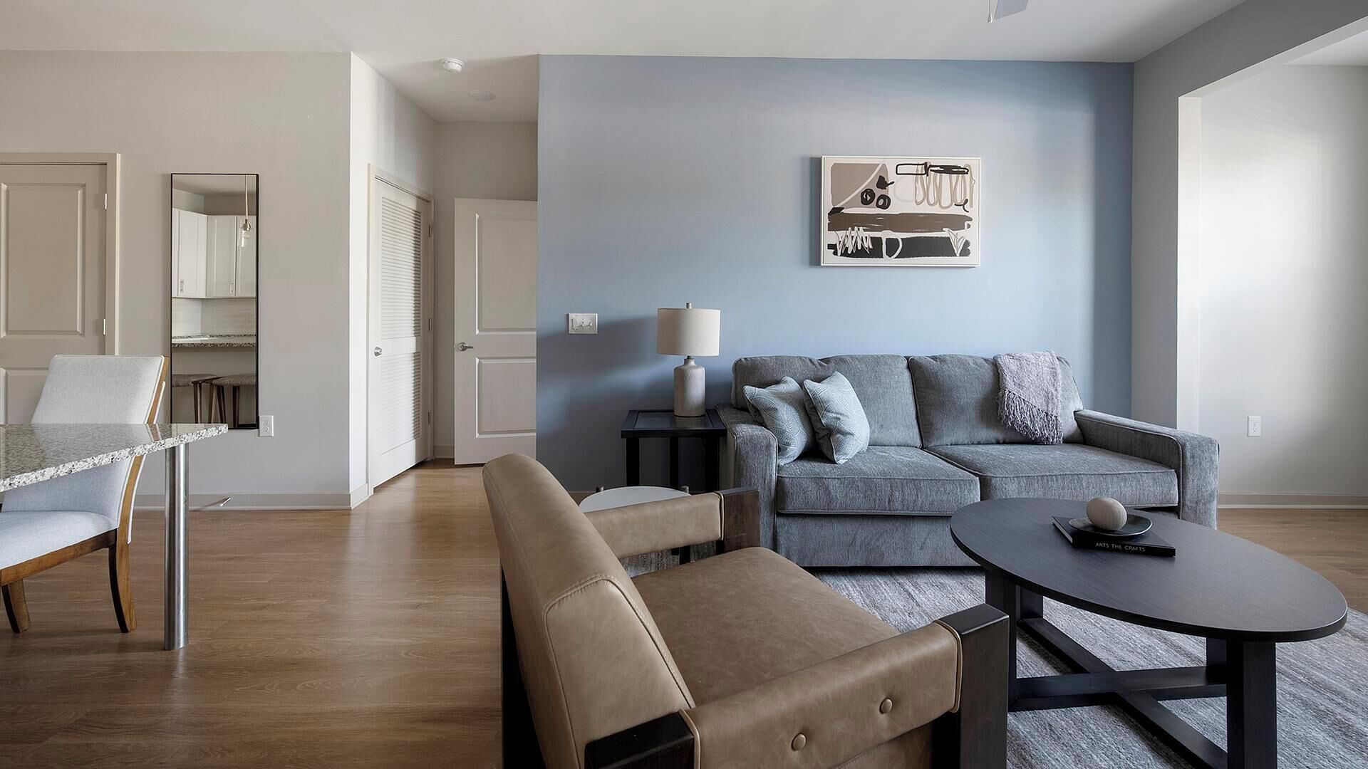 Ein modernes Wohnzimmer mit einem grauen Sofa, einem runden Couchtisch und einem beigefarbenen Sessel. Ein Beistelltisch mit Lampe und ein gerahmtes Bild hängen an der Wand.