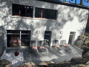 Ein modernes Gebäude mit großen Fenstern, einer Terrasse mit Lounge-Möbeln und einem gepflasterten Weg.