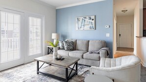 Una sala de estar con un sofá gris, una mesita de centro y un sillón blanco. Hay un cuadro en la pared y una ventana con persianas.