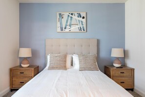 A bedroom with a tufted headboard, two bedside tables with lamps, a bed with white bedding, and a framed abstract artwork on the wall.