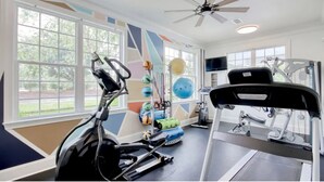 Un gimnasio con equipo de ejercicio, ventanas amplias y un ventilador de techo.