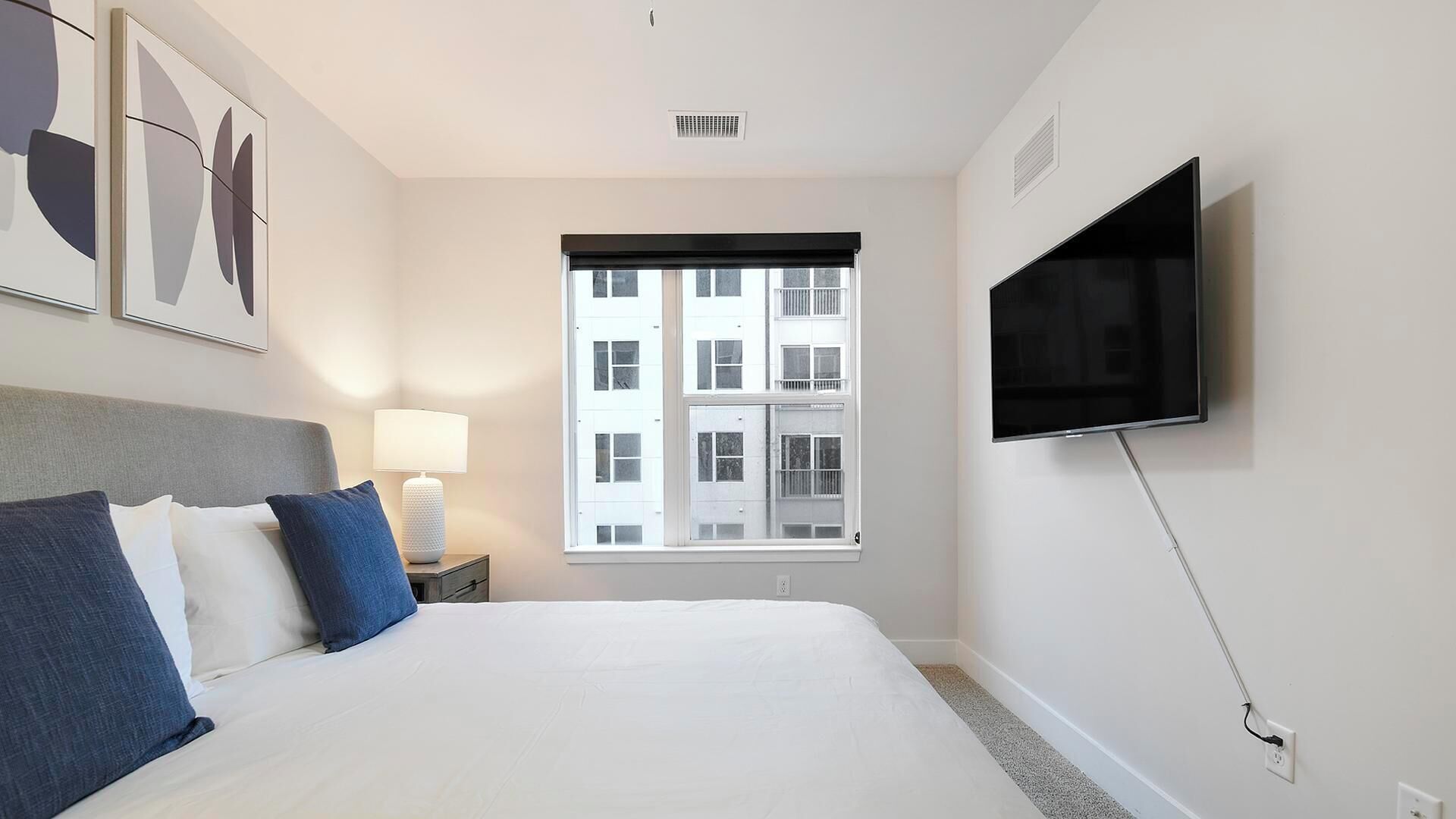 A hotel room with a bed, a TV mounted on the wall, a window with blinds, and artwork on the wall.