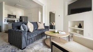 Ett modernt vardagsrum med en grå soffa, ett runt soffbord i trä och en beige fåtölj. I bakgrunden syns ett kök med vita skåp och vitvaror.