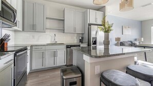 Una cocina moderna con armarios grises, un refrigerador de acero inoxidable, horno empotrado y encimera de granito. Hay un taburete pequeño y una barra con dos taburetes.