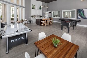 Cozinha moderna com armários brancos, sala de jantar com mesa e cadeiras de madeira, mesa de futebol e uma janela grande que traz luz natural.