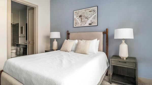 Un dormitorio ordenado con una cama, dos mesitas de noche con lámparas, una obra enmarcada en la pared y un baño visible a través de una puerta abierta.