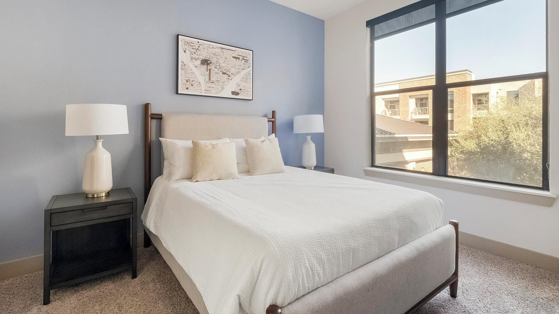 Un dormitorio con una cama grande, dos mesitas de noche con lámparas, un mapa enmarcado en la pared y una ventana que ofrece vistas a edificios.
