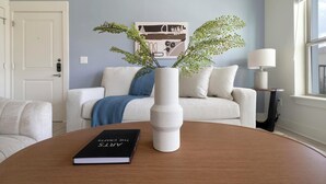 Uma sala de estar com um sofá branco, uma mesa de centro redonda de madeira, um livro intitulado 'THE CREATES' e um vaso branco com folhagem verde.