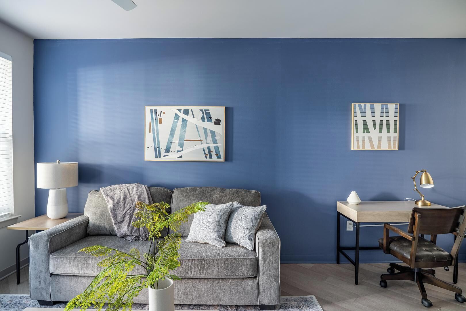A modern living room with a blue accent wall, a grey sofa, a small side table with a lamp, a desk with a chair, and two abstract paintings.