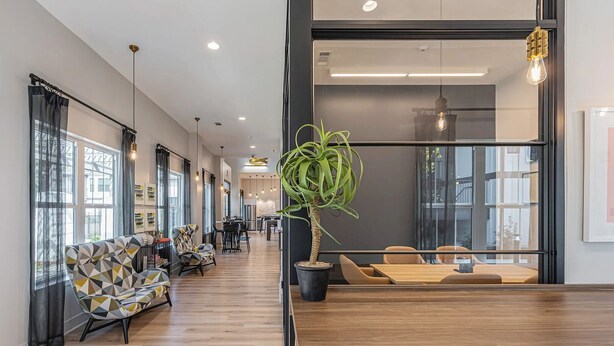 A modern hotel lobby with a long corridor, seating area with patterned sofas, a central potted plant, and a dining area with wooden tables and chairs.