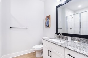 A modern bathroom with a white toilet, a granite countertop sink, a large mirror, and a shower area. - Landing | Beautiful 1BD, Game Room, Pool (Freeport)