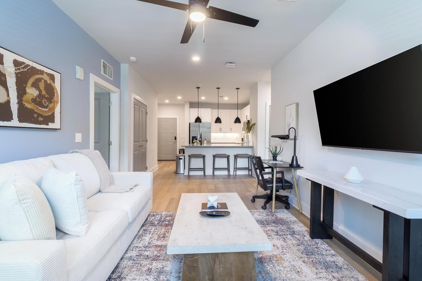 A modern living room with a white sofa, a coffee table, a TV mounted on the wall, and a kitchen in the background.