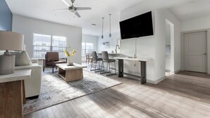 Uma sala moderna com sofá, mesa de centro e cadeiras, área de jantar com mesa e cadeiras, cozinha com televisão e uma janela grande com persianas.