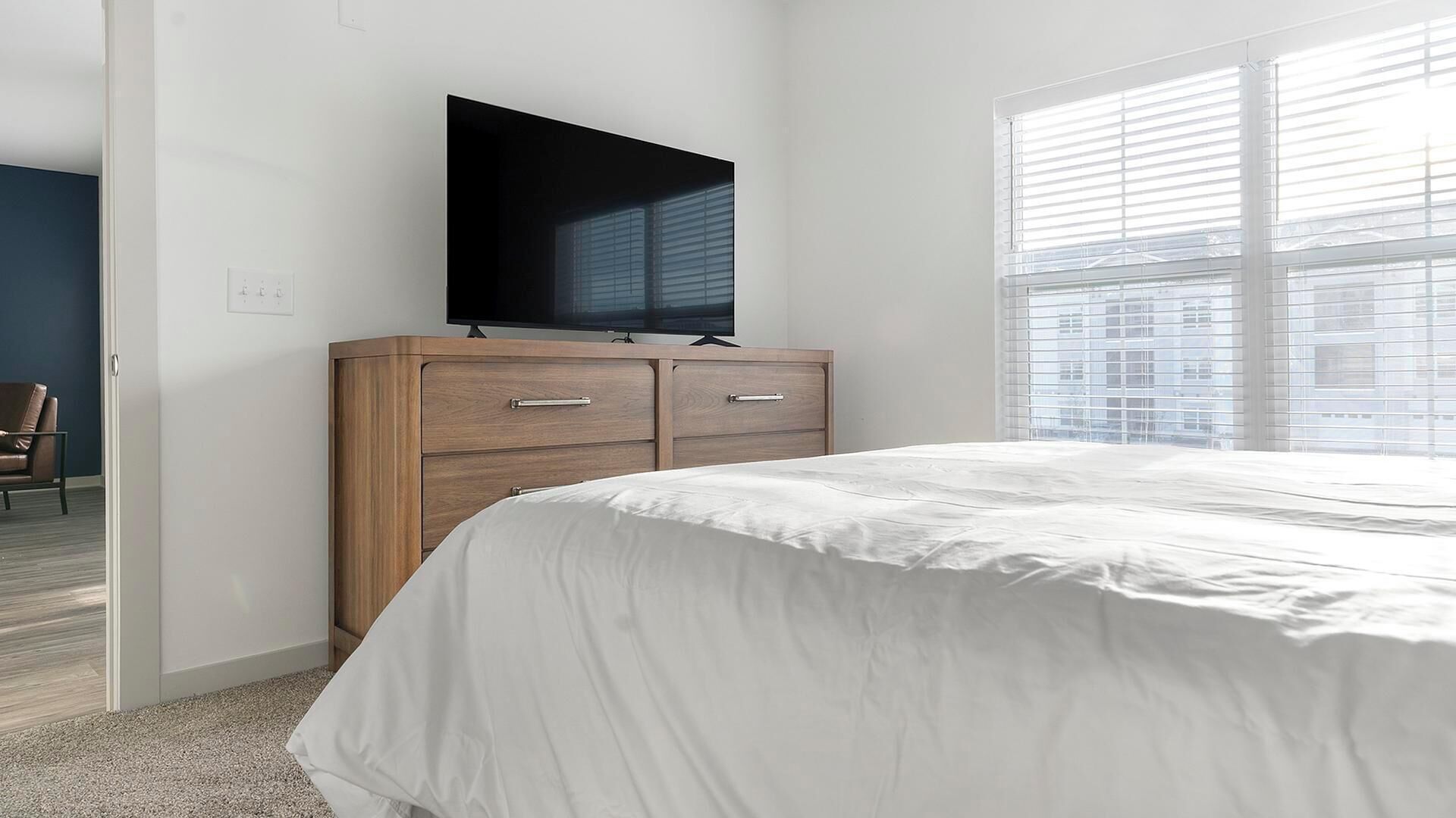 A bedroom with a bed, a wooden dresser with a flat-screen TV, and a large window with blinds.